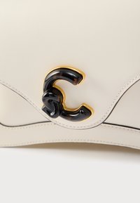 Crèmekleurige leren tas met een gebogen vorm, voorzien van een zwart-gouden logo-accent en fijne stiksels langs de randen. Soepele textuur.