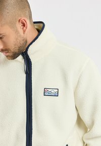 Fleece-Jacke in Creme mit einem dunkelblauen Kragen, durchgehendem Reißverschluss und einem Berg-Logo-Patch auf der linken Brust. Weicher Stoff, lässige Passform.