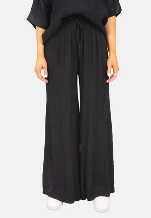 Black pleated wide-leg trousers with an elastic waistband and drawstring. Paired with white sneakers, emphasizing the relaxed fit.