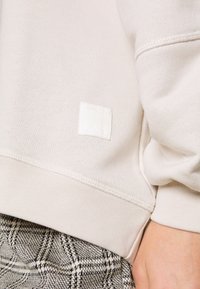 Gros plan d’une personne portant un sweat-shirt crème avec un petit patch logo carré et un pantalon à carreaux noir et blanc.