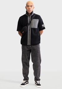 Veste en polaire noire avec des accents gris et une poche poitrine, associée à un jean gris foncé et des sneakers noires avec des lacets blancs.
