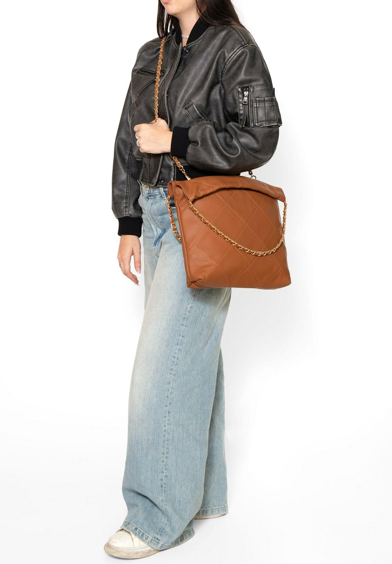 Borsa a tracolla marrone trapuntata con catena, forma rettangolare grande, texture morbida e chiusura a roll-top, indossata sopra jeans azzurri e giacca nera.