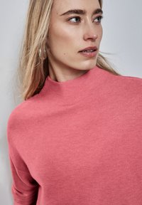 Pull rose à col montant en coton. Doté de manches longues et d'une finition texturée. Le visage du modèle est partiellement visible, avec des cheveux mi-longs.