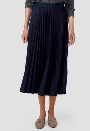 Femme portant une jupe midi plissée bleu marine, un top à manches longues imprimé et des mocassins taupe avec des détails dorés.