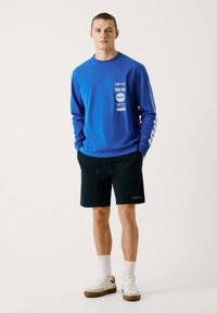 Blå sweatshirt med grafiske prints, parret med sorte shorts. Modellen har hvide sneakers og ankelsokker på. Enkel design, afslappet pasform.