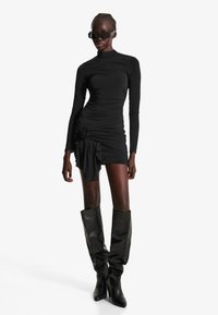 Črna obleka z dolgimi rokavi z gubanim teksturo, ki ima cvetlični poudarek na strani. V kombinaciji z črnimi usnjenimi škornji do kolen.