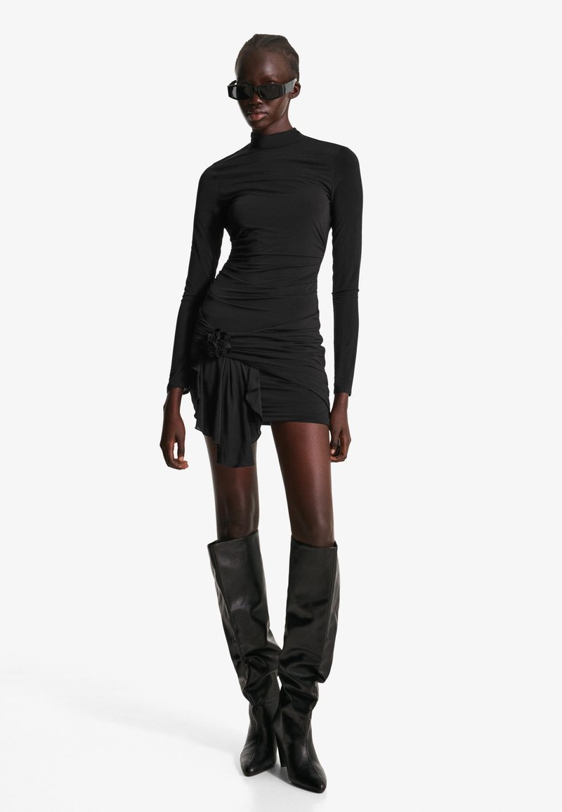 Črna obleka z dolgimi rokavi z gubanim teksturo, ki ima cvetlični poudarek na strani. V kombinaciji z črnimi usnjenimi škornji do kolen.