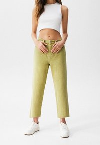 Jean taille haute vert avec coupe droite et ourlets effilochés, associé à un crop top blanc et des baskets blanches. Texture lisse.