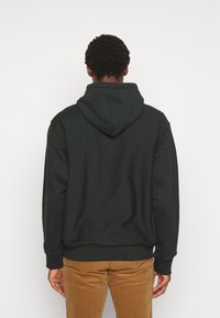 Felpa verde scuro con cappuccio regolabile, maniche lunghe e polsini a coste. Tessuto morbido e vestibilità comoda, abbinata a pantaloni in corduroy color sabbia.
