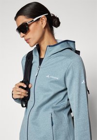 Casaco de fleece azul claro com fecho éclair à frente, capuz e bolsos laterais. Apresenta um logotipo no peito e uma alça ajustável de um lado.