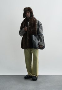 Hnedá vesta z umelého kožušiny cez sivú koženú bundu, pruhované tričko, zelené široké nohavice a čierne topánky. V ľavej ruke držiaca čiernu tašku.