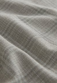 Tessuto strutturato in grigio chiaro con un sottile motivo a quadretti, che mostra increspature e pieghe, evidenziando la sua struttura tessuta e la superficie liscia.