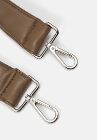 Extrémités de la sangle d'un sac en cuir marron avec crochets mousquetons pivotants en métal argenté sur un fond gris clair.