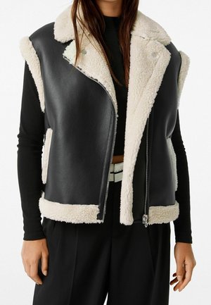 Femme portant un gilet en cuir noir sans manches doublé de shearling blanc, un haut noir à manches longues et un pantalon noir avec une ceinture blanche.