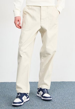 Beige bomullbyxor med avslappnad passform, med två framfickor och slät front. Kombineras med marinblå och vita sneakers.