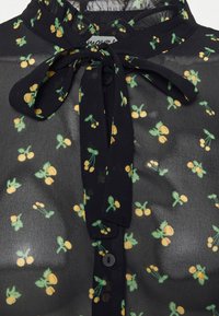 Blusa preta transparente com um pequeno padrão de cerejas amarelas e verdes. Inclui um laço atado no pescoço e um fecho frontal com botões.