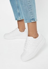 Valged nahast spordijalatsid, millel on sile tekstuur, paeltega disain, külgedel augulised detailid ja paks reljeefne tall. Denim-püksid, mis ulatuvad üle pahkluude.