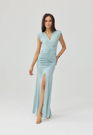 Kobieta w jasnoniebieskiej sukni ze zebraniem i rozcięciem do uda, stojąca z jedną ręką na biodrze, ubrana w przeźroczyste sandały na jasnym tle.