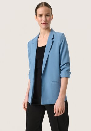 Světle modrý blazer vyrobený z hladké látky, s jednořadým zapínáním, klopovými límci a dvěma předními kapsami. Převlečený přes černý top.