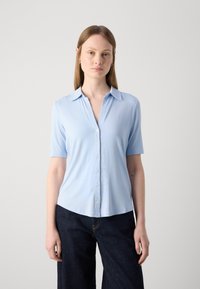 Chemise à manches courtes bleu clair à boutons en tissu lisse. Présente un col et un ourlet légèrement arrondi, associée à un pantalon en denim foncé.