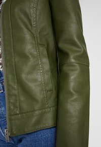 Giacca in pelle verde oliva con texture liscia, dotata di tasca laterale, chiusura con zip e maniche strutturate. Jeans in denim visibili sotto.