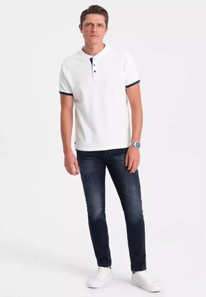 Polo bianca con dettagli blu navy e patta a tre bottoni, abbinata a jeans slim-fit blu scuro e scarpe da ginnastica bianche.