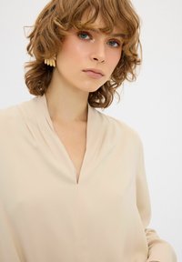 Beige Bluse mit V-Ausschnitt, aus glattem Stoff gefertigt. Verfügt über lange Ärmel und feine Drapierungen an den Schultern. Goldene Ohrringe als Akzente.