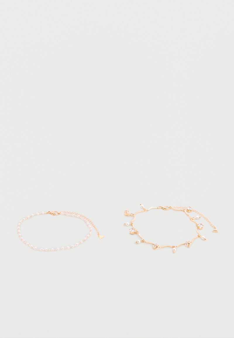 Deux bracelets délicats en chaîne d'or, l'un avec de petites perles, l'autre avec des breloques en perle et cristal, posés à plat sur une surface pâle.