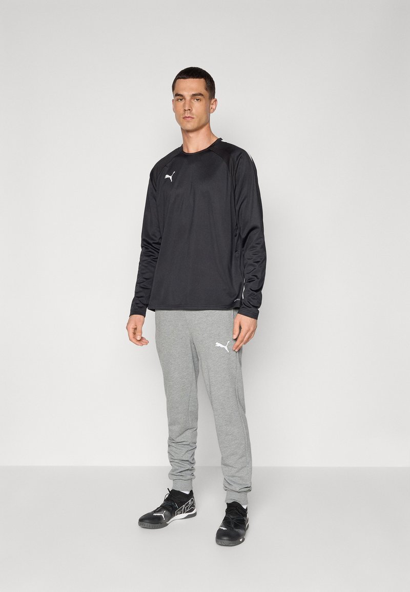 Zwart sport shirt met lange mouwen en een subtiele logo, gecombineerd met grijze joggingbroek met opgerolde enkels en een logo aan de zijkant. Zwarte sportschoenen.