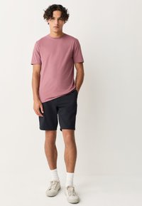 Lyserød bomuld t-shirt med korte ærmer, kombineret med marineblå shorts. Hvide ankelsokker og beige sneakers fuldender outfittet.