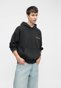Czarna bluza z kapturem z miękkiego materiału, z kieszenią z przodu i pomarańczowym logo. Połączona z jasnoniebieskimi dżinsami, tworząc casualowy look.