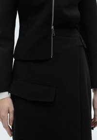 Giacca nera su misura con chiusura a zip e gonna coordinata con una grande tasca laterale. Tessuto liscio e strutturato con un design minimalista.