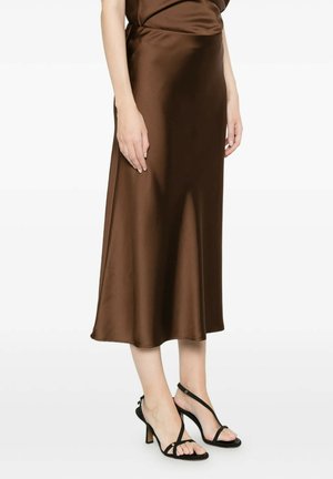 Donna che indossa una gonna midi in raso marrone abbinata a sandali neri con tacco alto e cinturini, in piedi davanti a uno sfondo bianco.