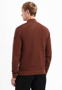 Homme de dos portant un pull marron texturé avec col, poignets et ourlet côtelés, associé à un pantalon noir sur fond blanc.