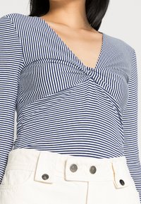 Långärmad topp i marinblå och vita ränder med en tvistad frontdetalj, kombinerad med krämfärgade hög midja-byxor med metallknappar.