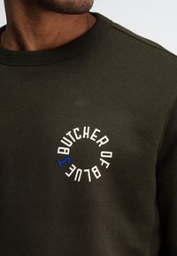 Olivengroene sweatshirt gemaakt van zachte stof, met een geribbelde halslijn en een ronde logo met "BUTCHER OF BLUE" in wit en blauw.