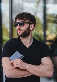 Černé tričko s výstřihem do V, které nosí muž držící šedý držák na karty. Černé sluneční brýle a světlý vous doplňují jeho ležérní vzhled. Přírodní pozadí.