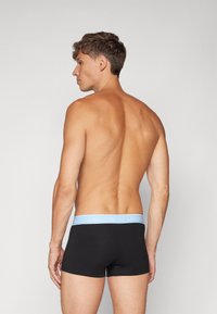 Svarte tettsittende boxershorts med en lyseblå Lacoste-logo på midjebåndet, vist fra en sidevinkel som fremhever det glatte stoffet og den tilpassede formen.