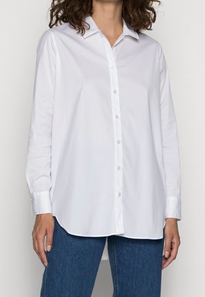 Femme portant une chemise blanche ample à manches longues avec col et un jean bleu, debout les bras détendus.