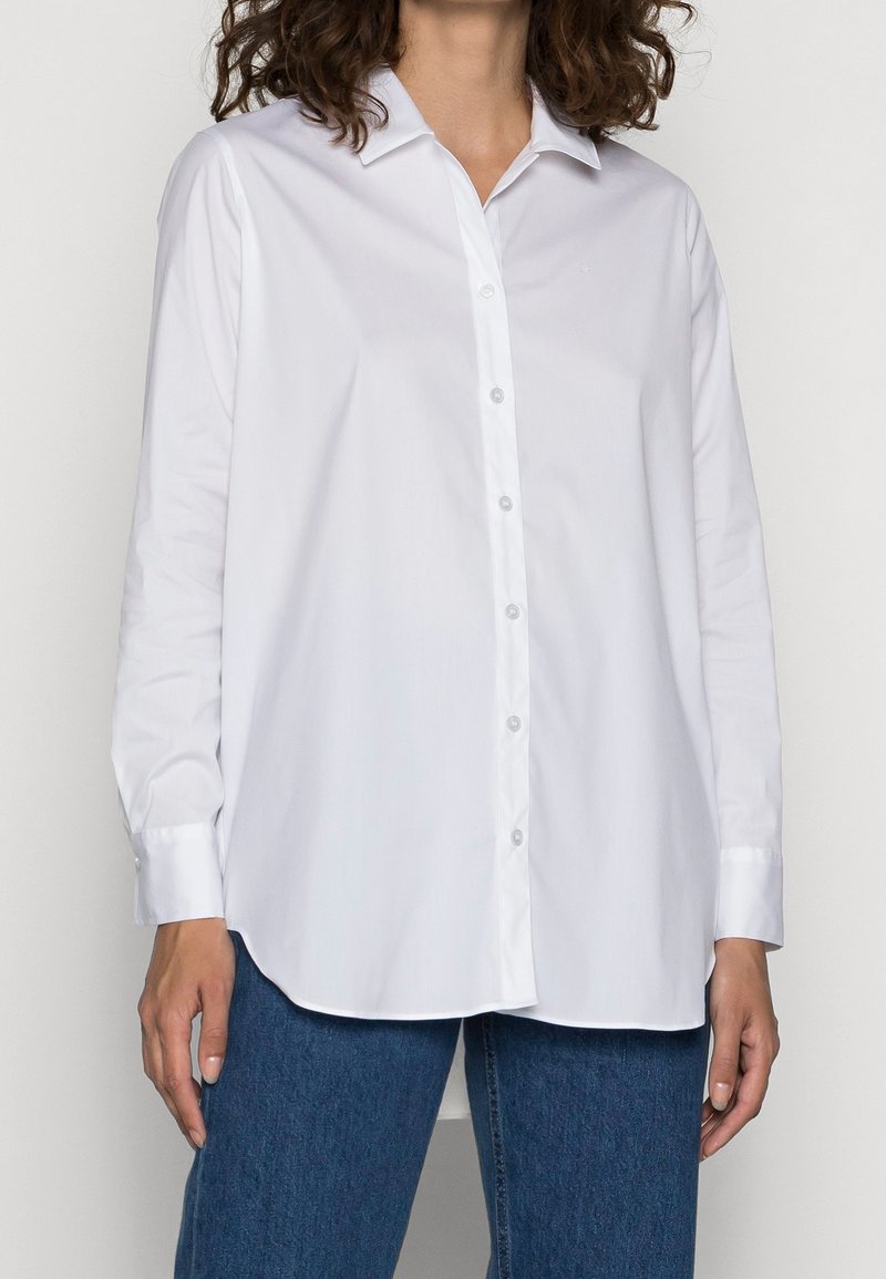Femme portant une chemise blanche ample à manches longues avec col et un jean bleu, debout les bras détendus.