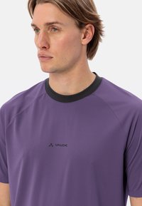 T-shirt de sport violet en tissu respirant, avec un col rond noir, des manches courtes et un logo imprimé sur la poitrine.