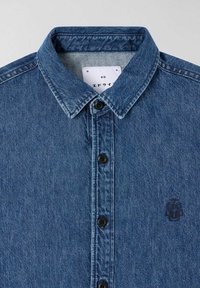 Camicia in denim di colore blu medio, con colletto button-down, bottoni neri e un sottile dettaglio del logo sul lato sinistro.