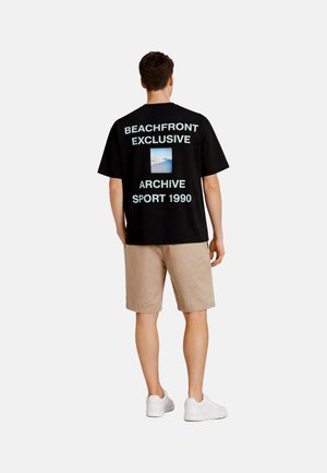 Man wearing black oversized t-shirt with "BEACHFRONT EXCLUSIVE ARCHIVE SPORT 1990" and a beach photo, beige shorts, and white sneakers.