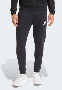 Μαύρο αθλητικό παντελόνι από λείο ύφασμα, με στενή εφαρμογή, ελαστικά μανσέτα και λευκό λογότυπο Adidas στο μηρό.