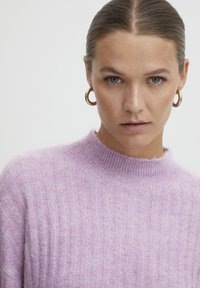 Maglione a coste di colore viola chiaro con collo alto, realizzato in materiale morbido. Il modello indossa semplici orecchini a cerchio e uno sfondo neutro.
