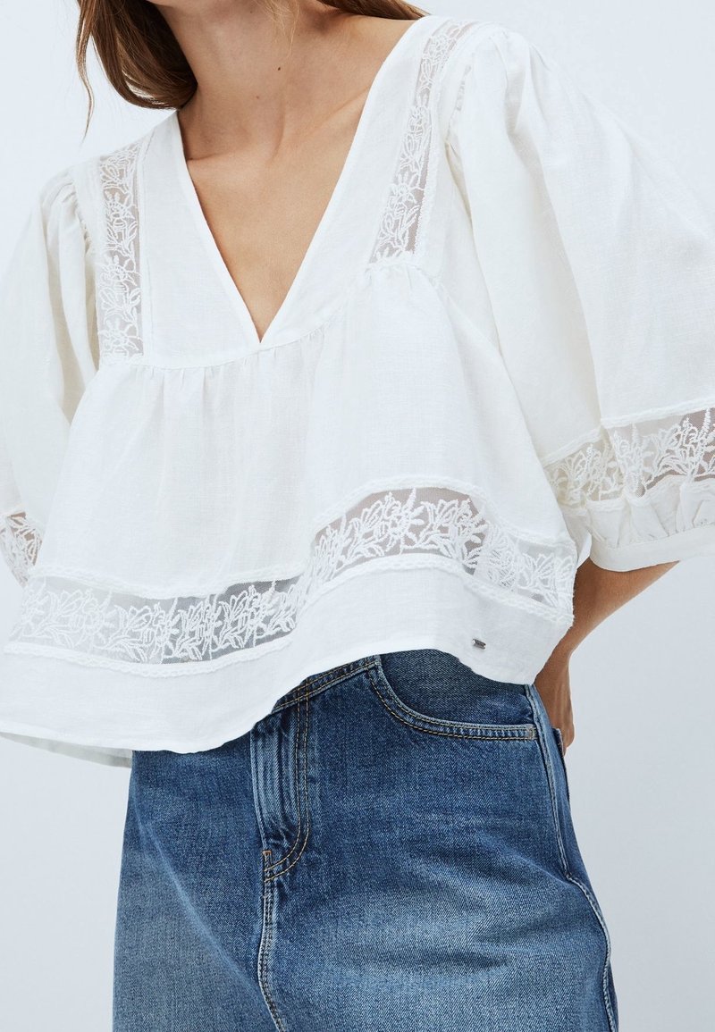 Vit blus med djup V-ringning, med spetsdetaljer och vida ärmar. Buren över blå jeans, vilket ger en lös och avslappnad passform.