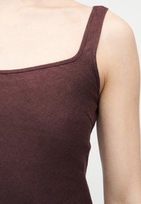 Κοντινό πλάνο ενός ατόμου που φοράει αμάνικο τοπ σκούρου καφέ με τετράγωνη λαιμόκοψη, εμφανίζοντας τους ώμους και την περιοχή πάνω από το στήθος.