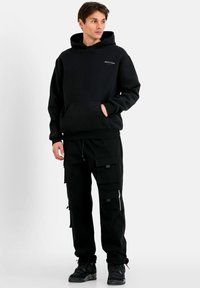 Schwarzer Hoodie mit Fronttasche und verstellbarem Kapuzenzug. Kombiniert mit schwarzen Cargo-Hosen, die mehrere Taschen und Reißverschlussdetails aufweisen.