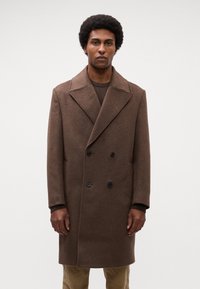Cappotto marrone doppiopetto con ampi revers, realizzato in tessuto strutturato. Presenta bottoni neri e una vestibilità rilassata, abbinato a un top lavorato a maglia.