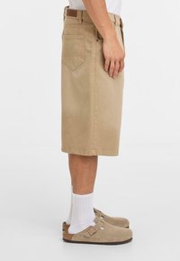 Beige katoenen shorts met een rechte pasvorm en knielengte, voorzien van zij- en achterzakken. Gepaard met witte sokken en tan klompen.